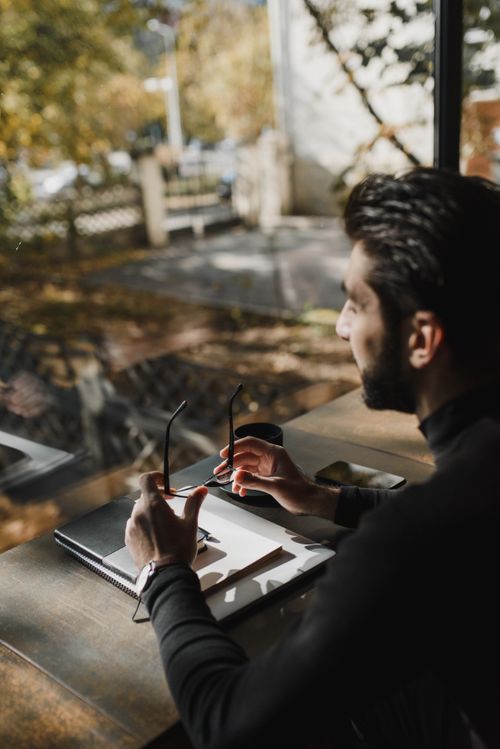 Free Man sitting indoors by a window, holding eyeglasses, in thoughtful mood. Stock Photo