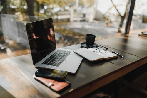 Free A serene office setup with a laptop, notebook, and coffee near a window, reflecting an autumn scene. Stock Photo