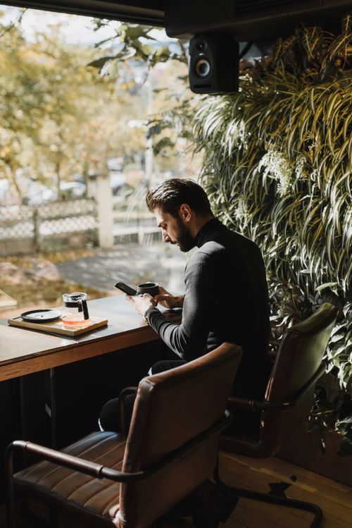 Free A man in a café sits by the window, focused on his phone, surrounded by sunlight and greenery. Stock Photo