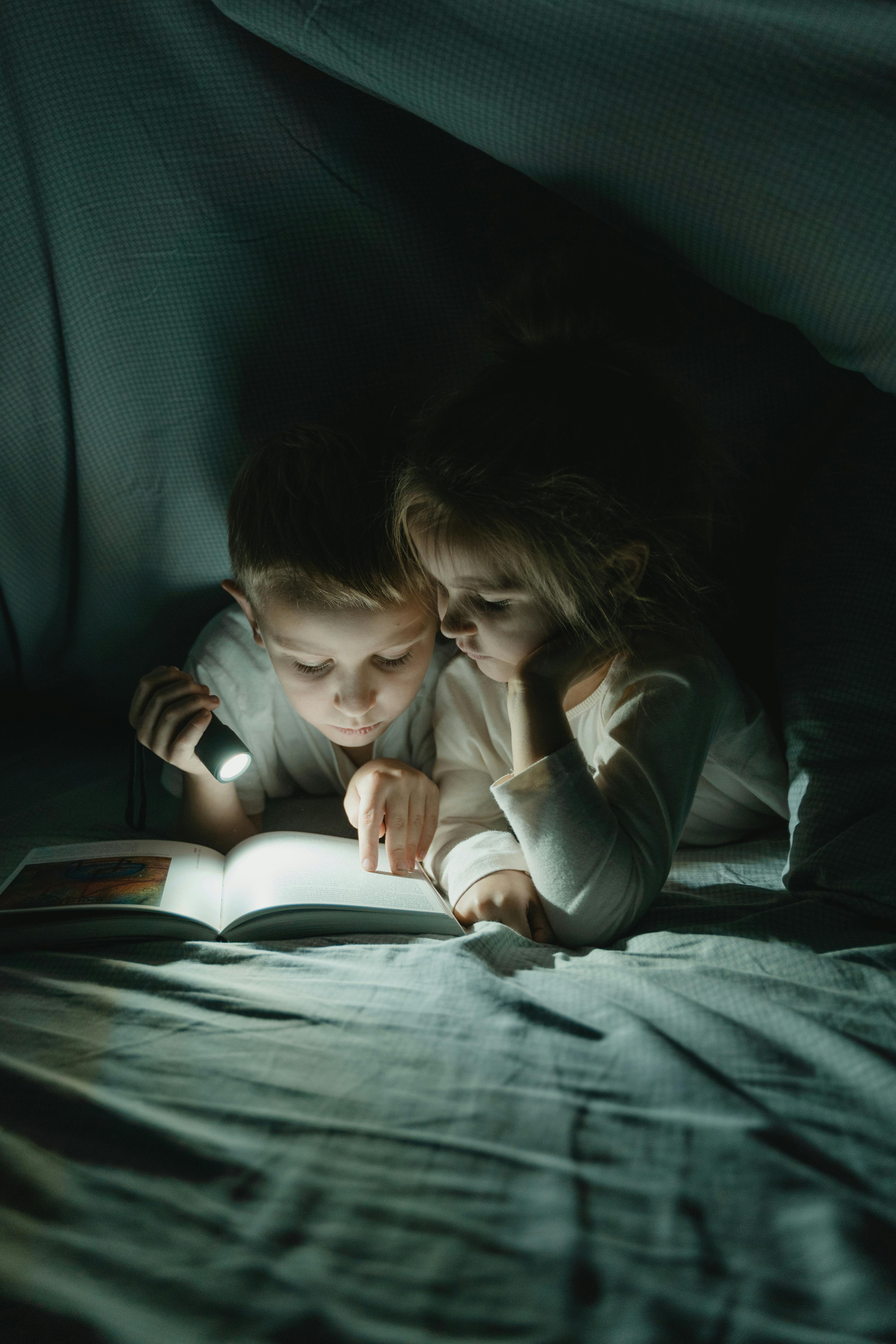 girl and boy reading book under blanket