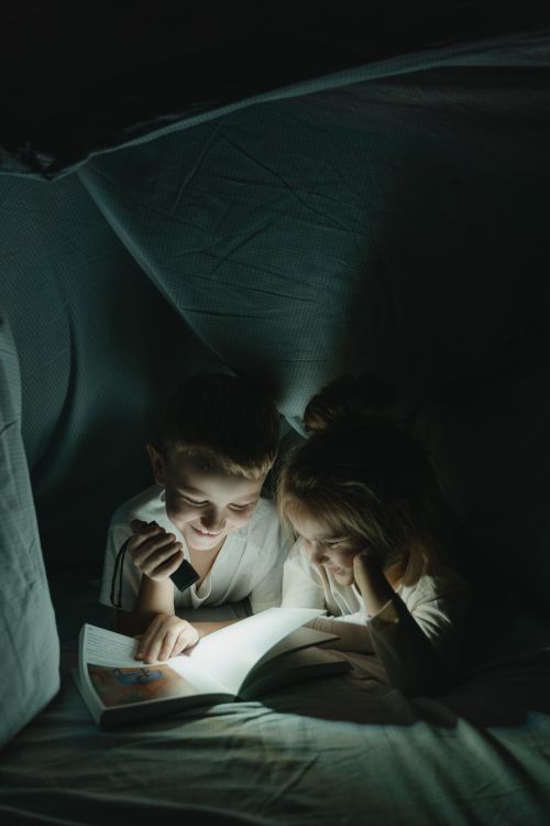 Free Two children enjoy reading a book under a cozy blanket fort, creating a magical nighttime moment. Stock Photo