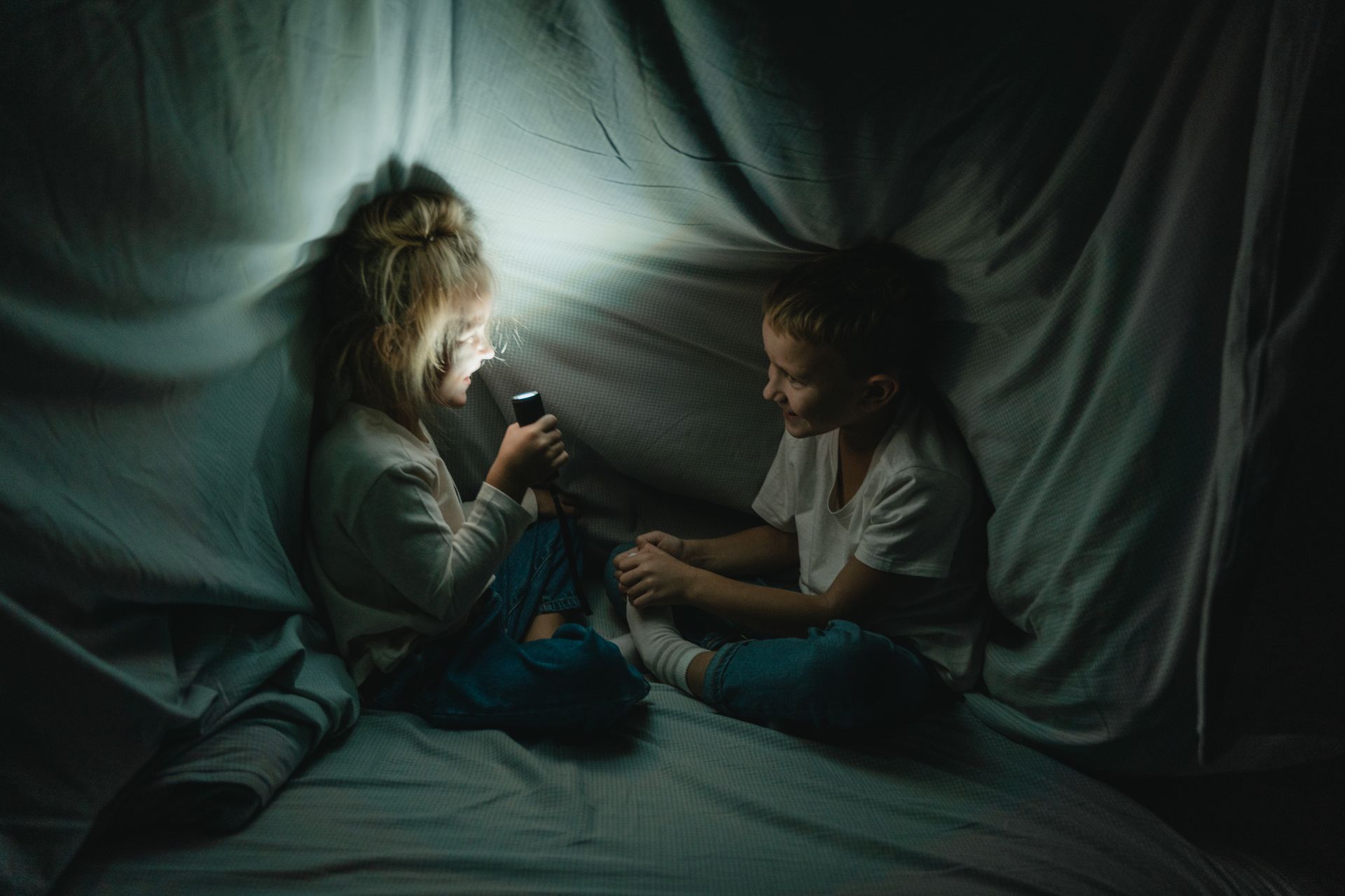 Two children enjoying a storytime under a blanket fort with a flashlight at night.