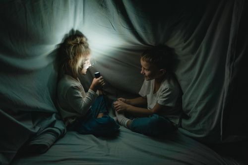 Free Two children enjoying a storytime under a blanket fort with a flashlight at night. Stock Photo
