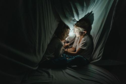 Free Two kids enjoy storytelling under a cozy blanket fort lit by a flashlight. Stock Photo
