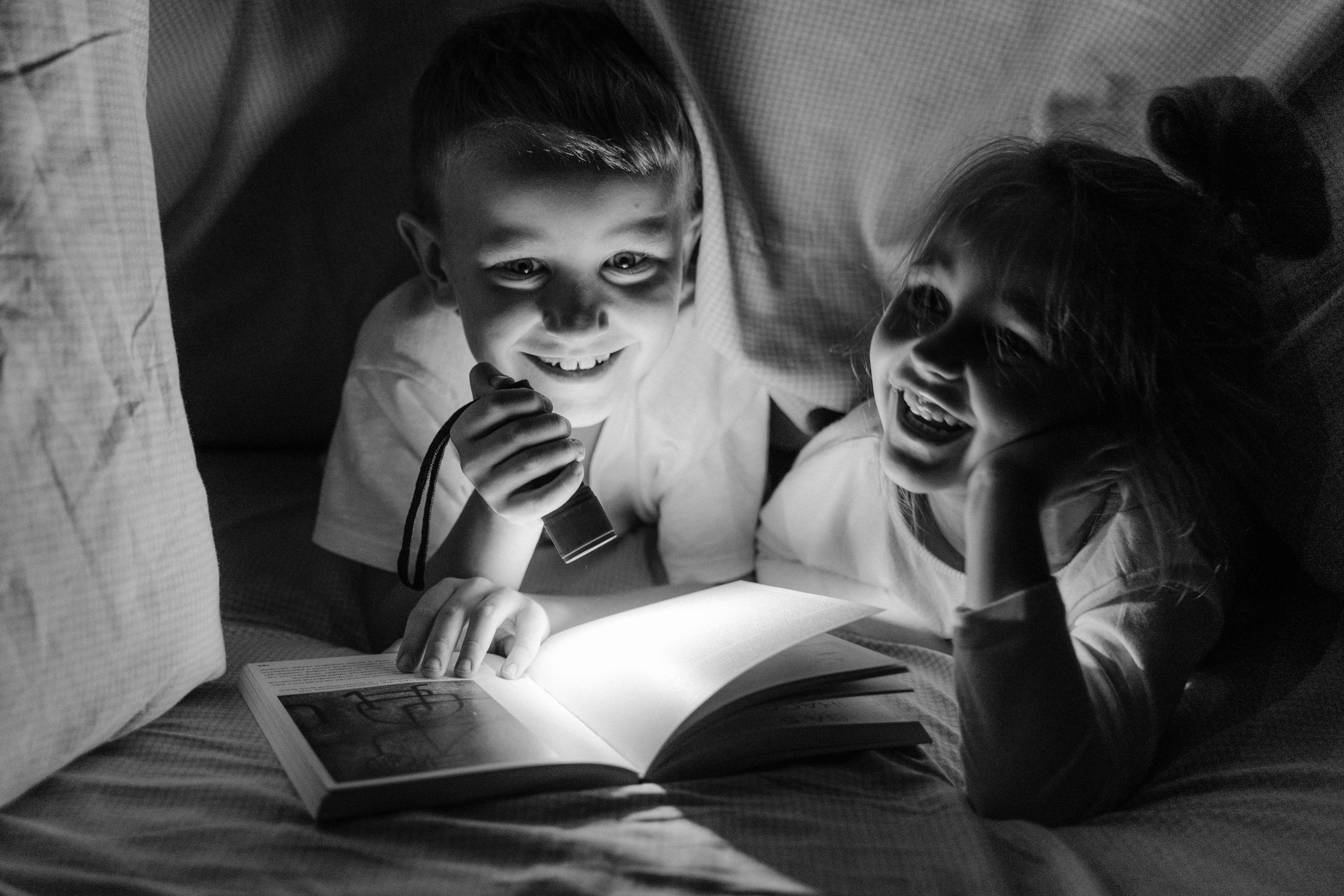 grayscale photo of girl and boy reading book under blanket and smiling