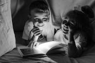 Grayscale Photo of Girl and Boy Reading Book Under Blanket and Smiling