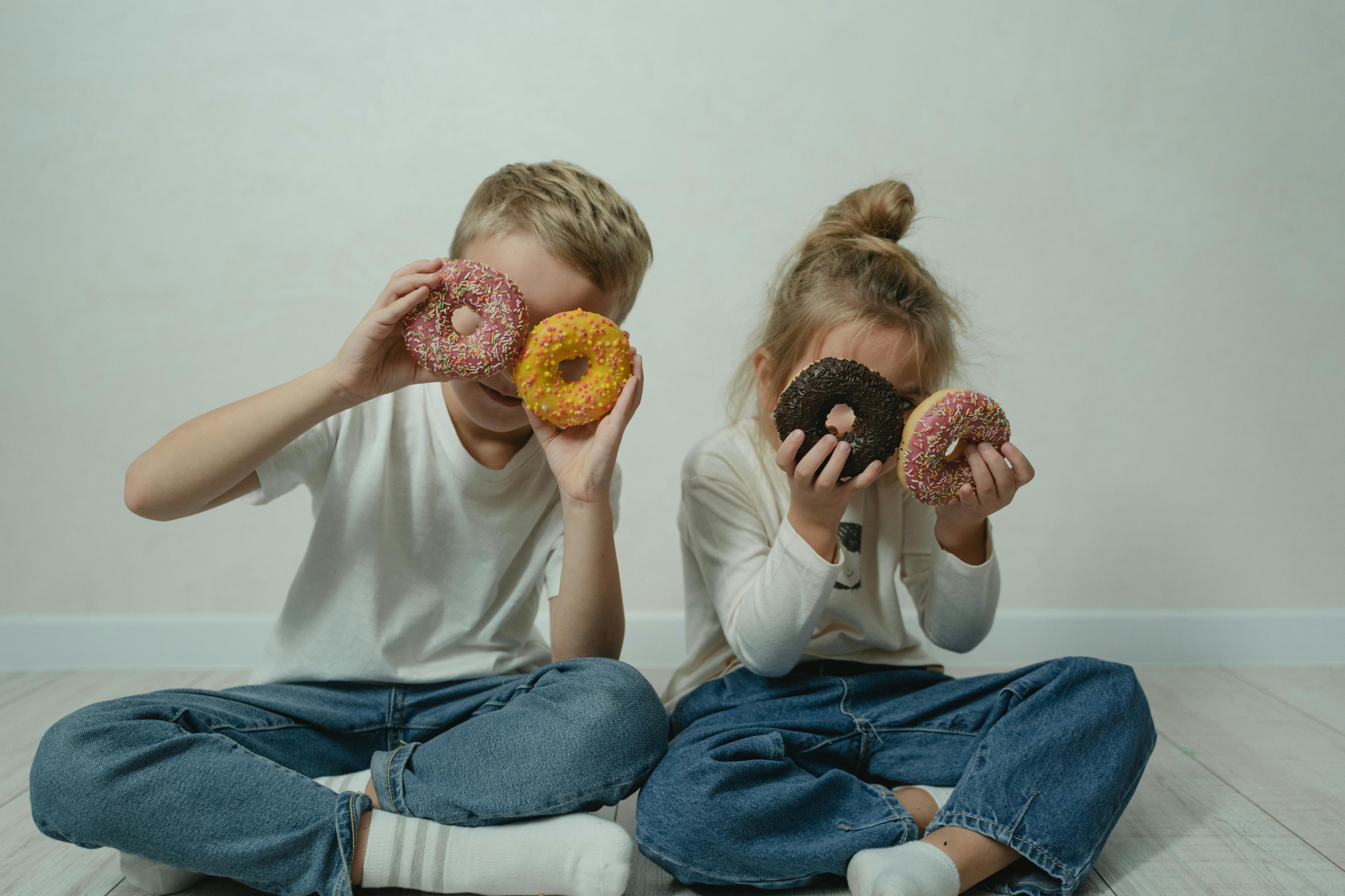 两个孩子坐在屋里，开玩笑地把撒了彩色糖霜的甜甜圈放在眼睛上