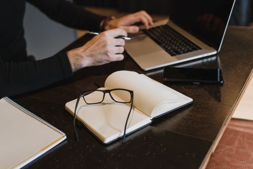 Free Professional setting with hands typing on laptop, notebook, and eyeglasses on table. Stock Photo