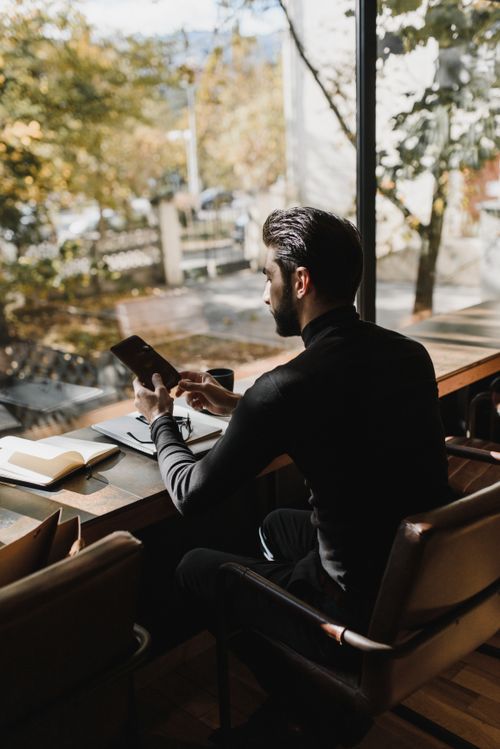 Free A young man sitting by a window in a cafe, using a smartphone next to a laptop with an autumn view outside. Stock Photo