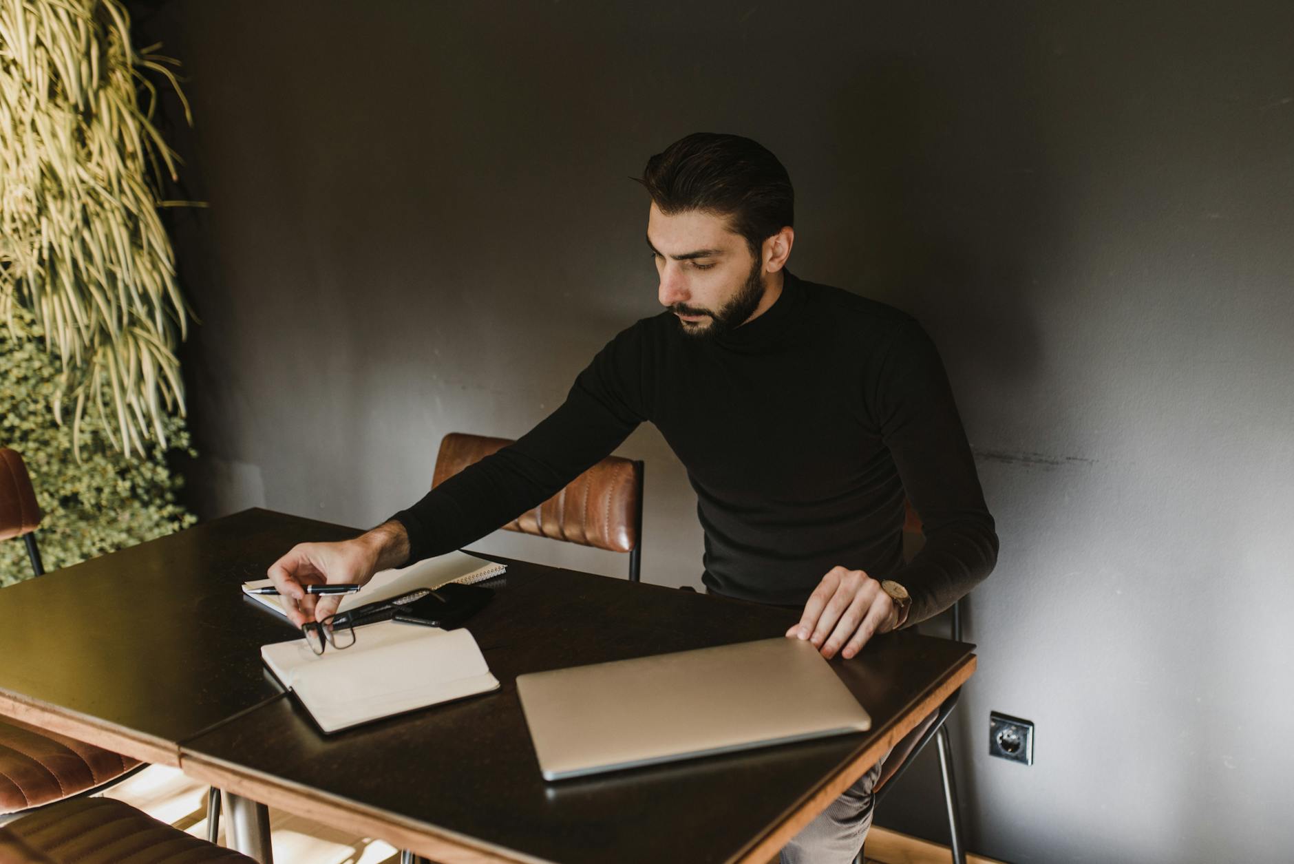 Adult man sitting at a desk working with a laptop, notebook, and pen indoors.