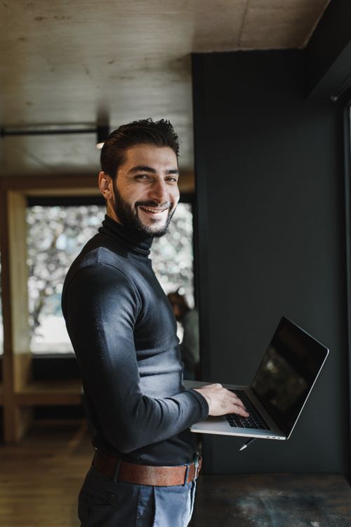 Free A man smiles while working on his laptop in a modern indoor setting. Stock Photo