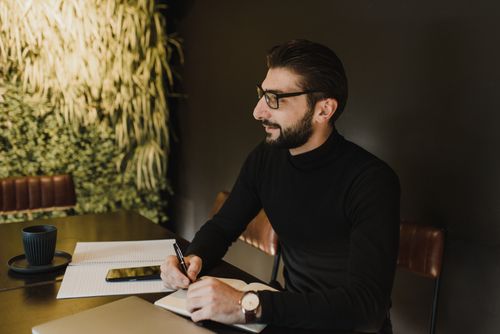 Free An adult man with eyeglasses writing in a notebook while sitting at a table indoors. Stock Photo