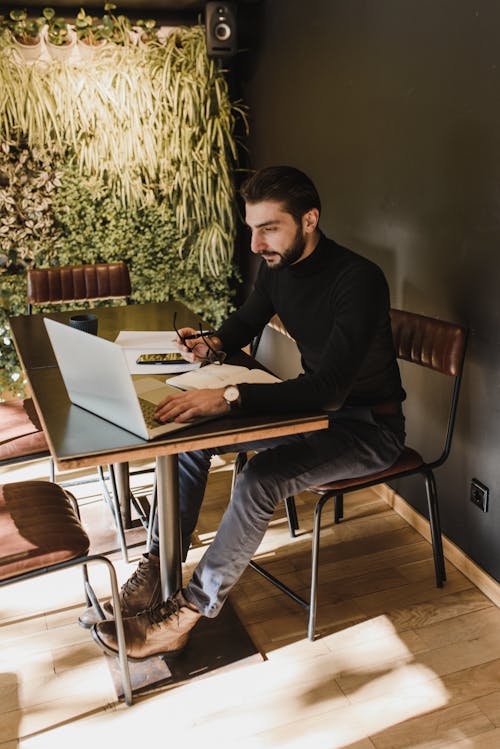 Side view full length young bearded male in casual clothes sitting at table with papers and browsing netbook while working in creative workplace