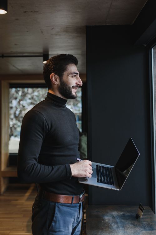 Free Man standing indoors with a laptop, smiling while looking outside. Perfect for business or lifestyle concepts. Stock Photo