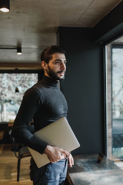 Free Side view of a man holding a laptop, looking out a large window in a modern setting. Stock Photo
