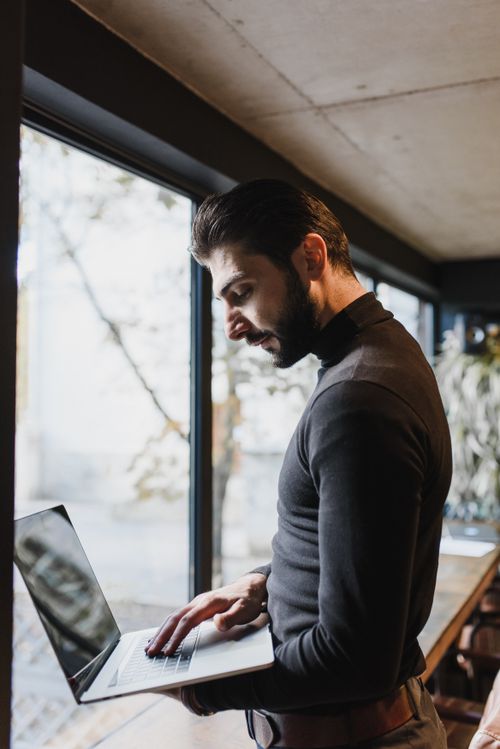 Free Adult man using a laptop indoors by a large window, focused on work. Stock Photo
