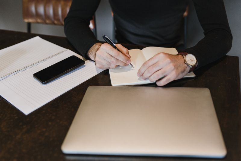 A person writing in a notebook with a laptop and smartphone on a table