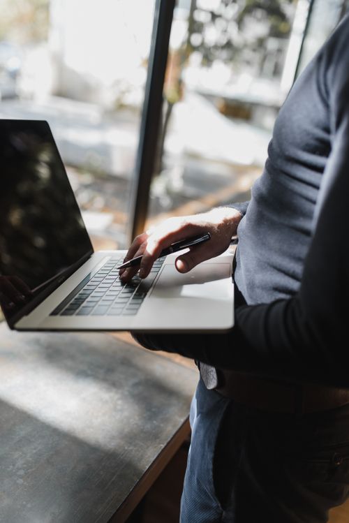 Free A man holding a laptop with one hand and pen in the other, near a sunlit window indoors. Stock Photo