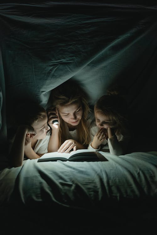 A Woman Reading a Book to her Children under a Blanket