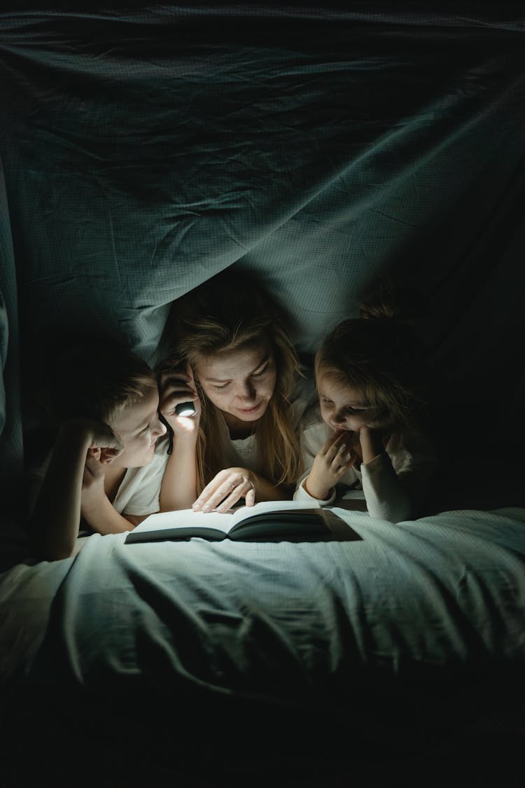 A Woman Reading A Book To Her Children Under A Blanket