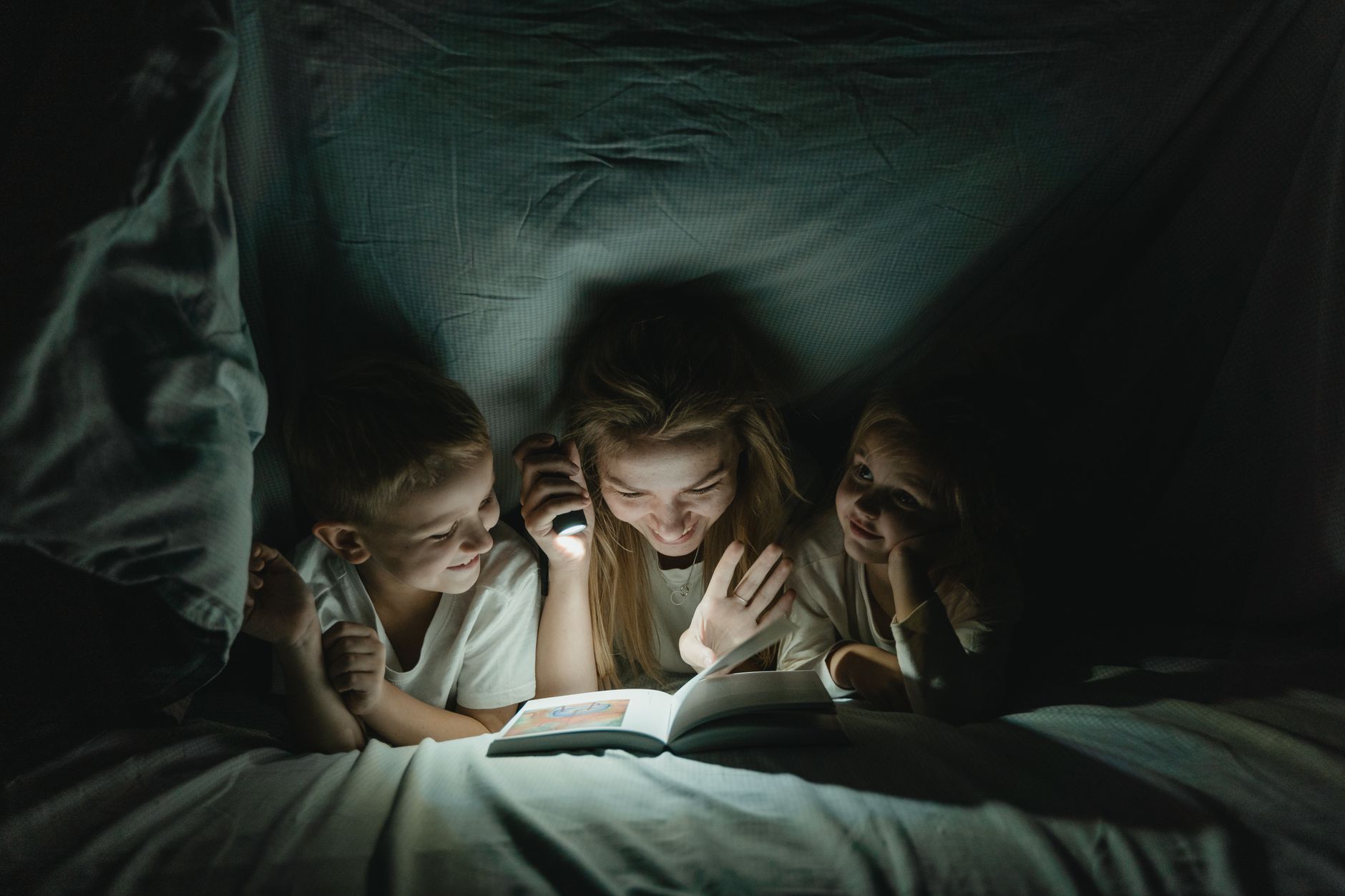 A mother shares a bedtime story with her kids under a cozy blanket, lit by a flashlight.
