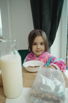 Adorable child in colorful pajamas eating cereal at a table with a milk bottle nearby.