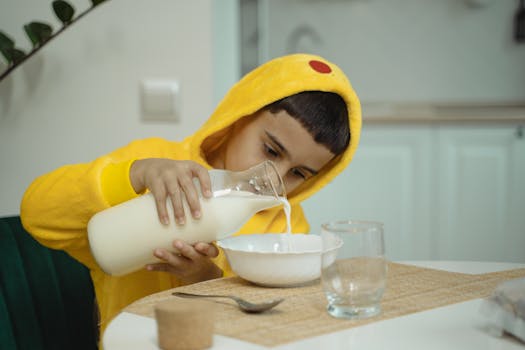 A child in a yellow hoodie carefully pouring milk into a bowl at the table.