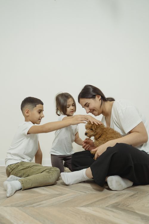 Free Mother and two children enjoying time with their puppy indoors, sitting on the wooden floor. Stock Photo