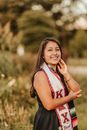 Smiling ethnic female student wearing sorority scarf looking at camera and touching face while standing near green plants on blurred background