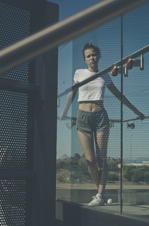 Low angle full length of serious young female in sportswear standing near glass building on balcony in sunny summer day under blue cloudless sky and looking at camera