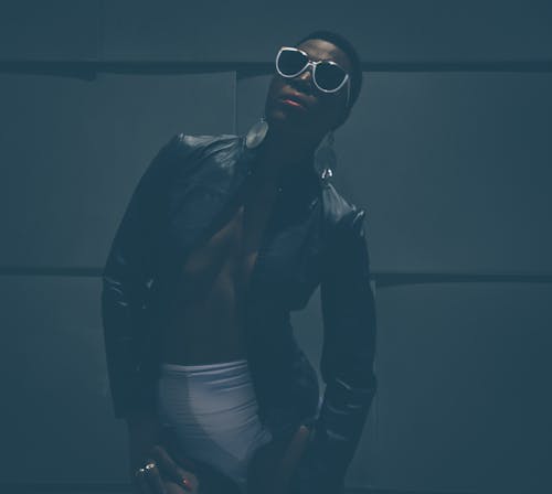 Confident black female model in trendy outfit and sunglasses and in earrings standing in dark near uneven wall