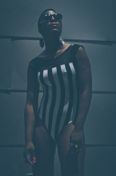 African American woman model in trendy swimsuit and sunglasses and with earrings standing near uneven wall in dark and looking up