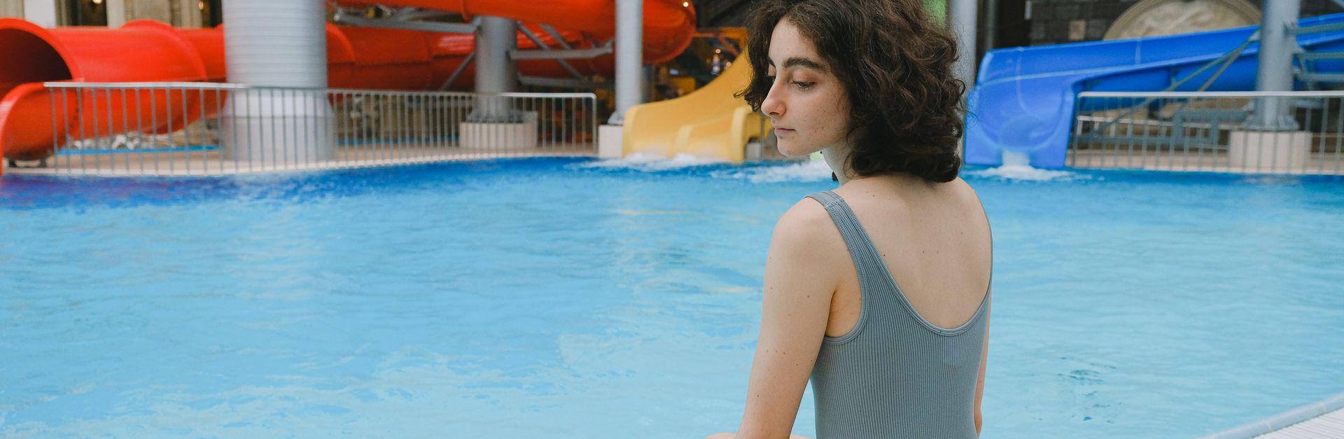 Young woman in a swimsuit sitting by an indoor swimming pool with colorful slides.