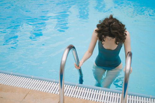 Free Young woman in a teal swimsuit using ladder to enter the pool on a sunny day. Stock Photo