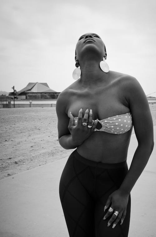 Black and white African American lady in bra standing on sandy shore and caressing chest in daytime