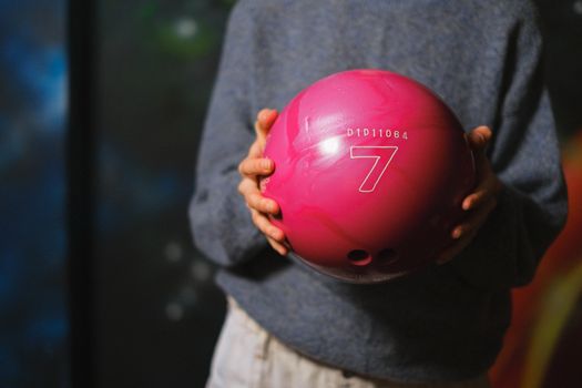 Child in a sweatshirt holding a pink bowling ball indoors. Perfect for sports and leisure themes.