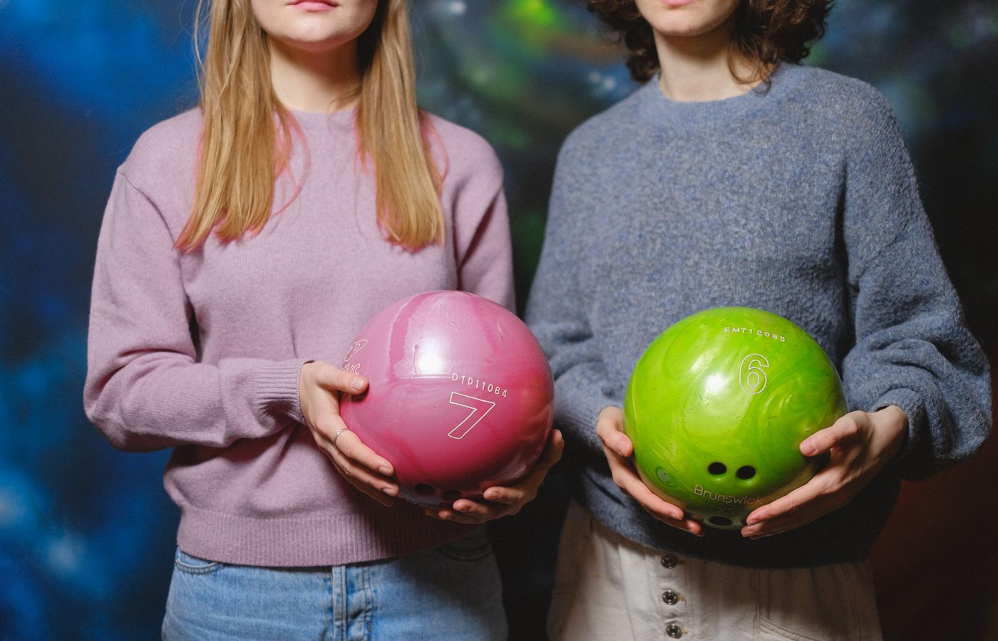 Two women holding pink and green bowling balls inside a bowling alley.