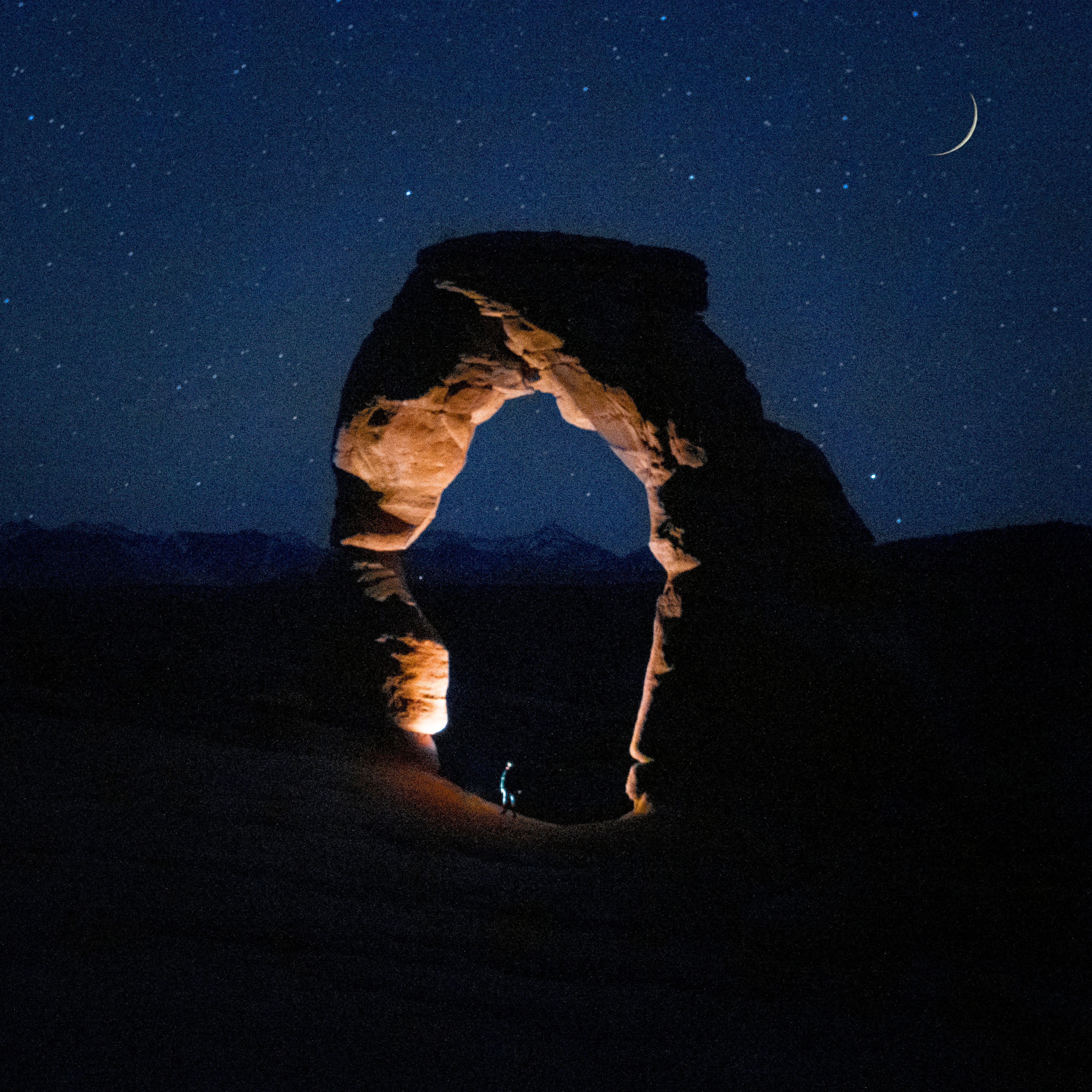 Nighttime view of Delicate Arch in Moab, Utah, under a starry sky with crescent moon.