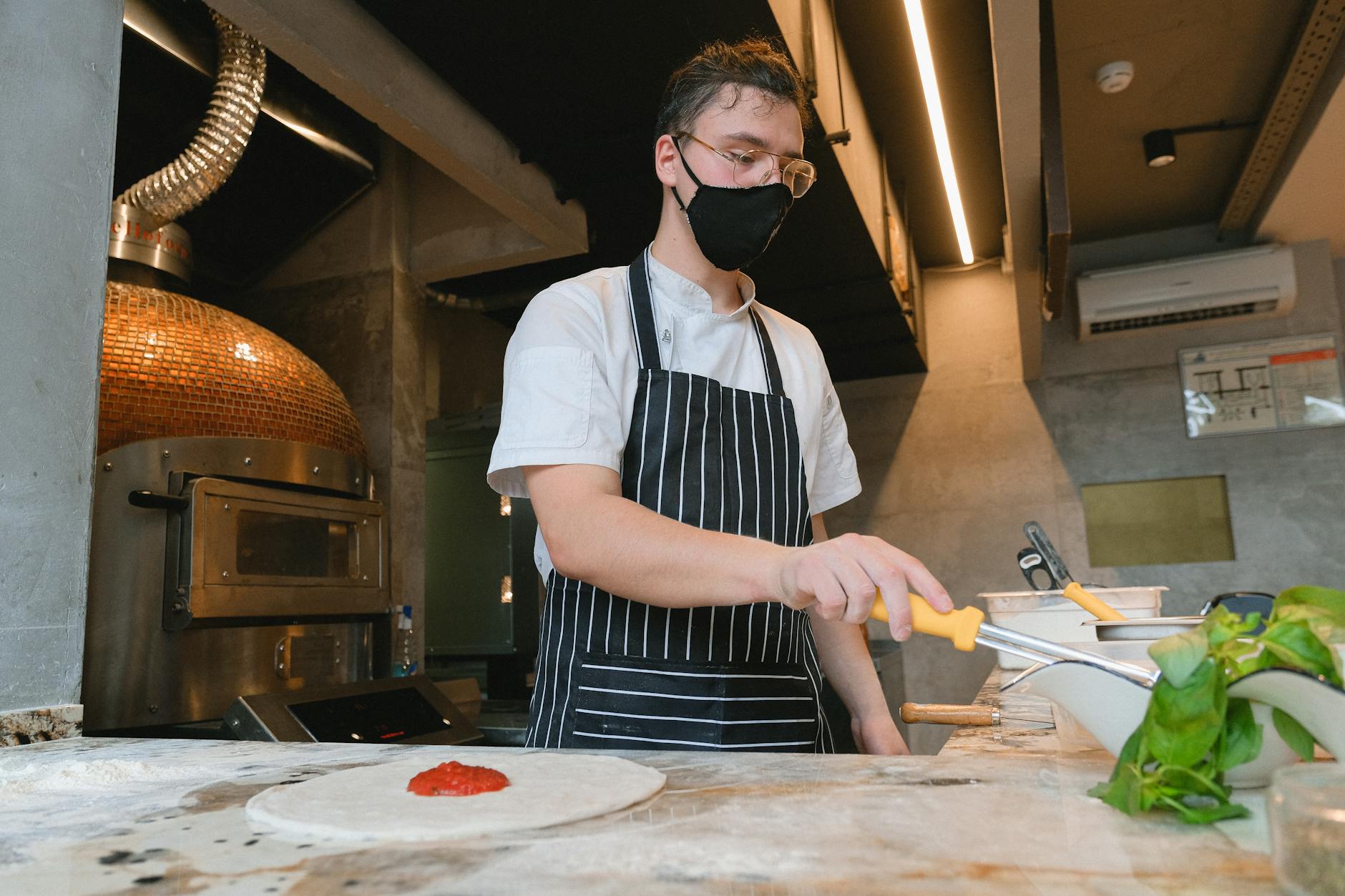 Chef in mask spreads toppings on pizza dough in a stylish modern kitchen.