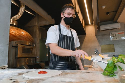 Free Chef in mask spreads toppings on pizza dough in a stylish modern kitchen. Stock Photo