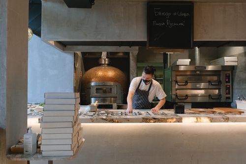 Free Masked chef preparing food in a modern pizzeria kitchen with ovens and pizza boxes. Stock Photo