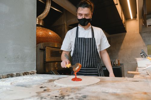 Free Chef wearing mask spreads tomato sauce on pizza base in kitchen. Stock Photo