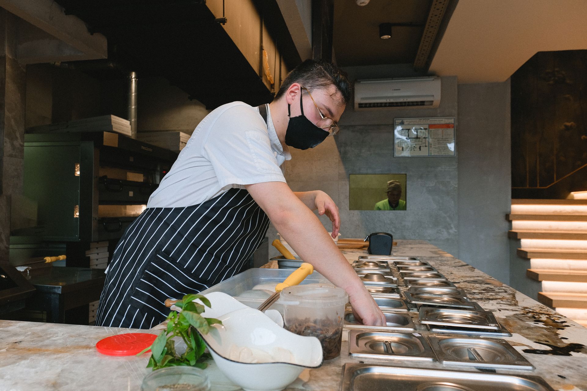 Masked chef in a modern kitchen preparing food with various ingredients, emphasizing safety and hygiene.