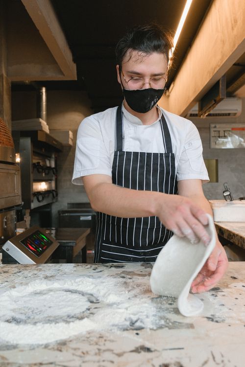 Free A chef in a face mask prepares dough on a floured surface in a restaurant kitchen. Stock Photo