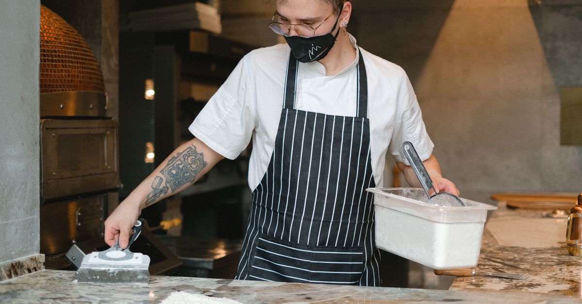 flour-dusted hands stretching pizza dough on a steel prep table beside a glowing deck oven, grated mozzarella tubs in the foreground