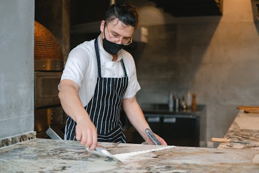 A chef wearing a face mask works on dough in a professional kitchen with marble counters.