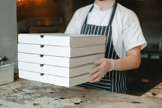 Pizza deliveryman carrying stacked boxes in a restaurant kitchen setting.