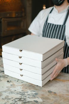 A pizza delivery worker holds stacked boxes in a restaurant kitchen, ready for delivery.