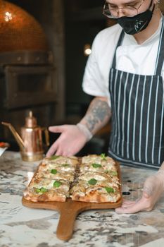Masked chef showcasing artisan pizza on wooden tray with fresh herbs.
