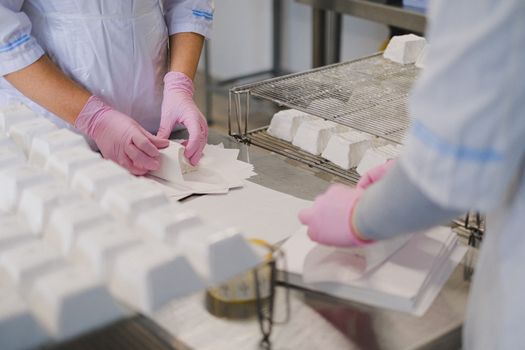 Workers in protective gear carefully wrapping cheese blocks by hand in a dairy factory setting.